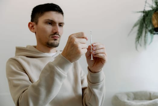 One young Caucasian handsome brunette man with a serious emotion takes medicine from a bottle with a syringe to treat his cat at home, standing against the background of a white wall, close-up side view.