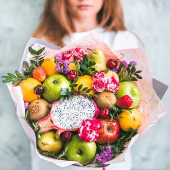 Fruit and berries bouquet. Eating bouquet in female hands. Apple, orange, strawberry, pear, kiwi, dragon fruit and flowers, eucalyptus. Shallow DOF