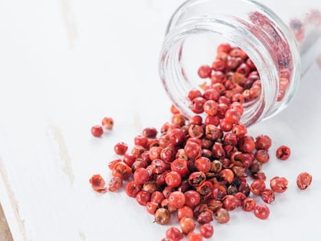 Dried pink pepper berries poured from glass jar on white wooden cutting board. Close up view of pink peppercorn poured on vintage wooden table. Copy space.