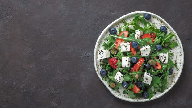 Salad with arugula, feta cheese and berries - strawberry, blueberry, in craft plate on black bacground. Summer salad idea and recipe for healthy vegetarian lunch, dinner. Top view. Copy space.