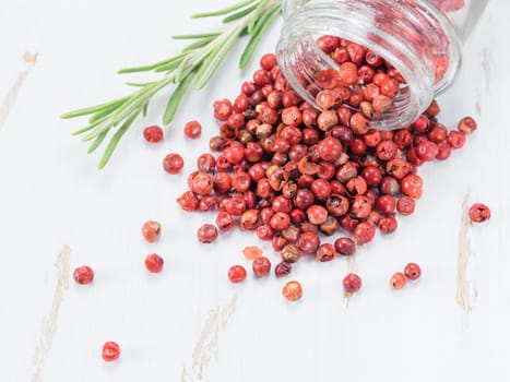 Dried pink pepper berries poured from glass jar on white wooden cutting board. Close up view of pink peppercorn and fresh rosemary on vintage wooden table. Copy space.