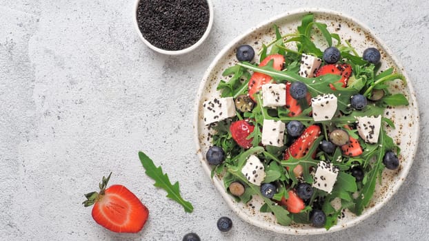 Salad with arugula, feta cheese and berries - strawberry, blueberry, in craft plate on gray cement bacground. Summer salad idea and recipe for healthy vegetarian lunch, dinner. Top view. Copy space.