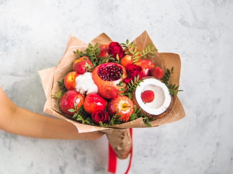 Fruit and berries bouquet. Eating bouquet in female hand on gray background. Pomegranate, strawberry, apples, coconut and roses flowers, eucalyptus. Shallow DOF. Copy space