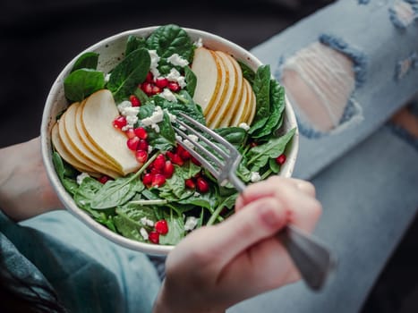 Woman in jeans holding vegan salad bowl with spinach, pear, pomegranate, cheese. Vegan breakfast, vegetarian food, diet concept. Girl in jeans holding fork with knees and hands visible