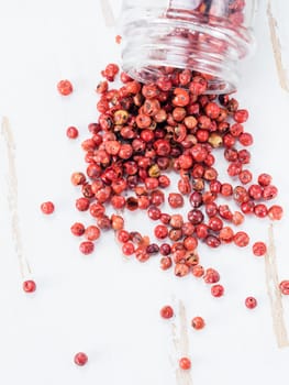 Dried pink pepper berries poured from glass jar on white wooden cutting board. Close up view of pink peppercorn poured on vintage wooden table. Copy space. Top view or flat-lay.