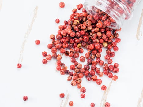 Dried pink pepper berries poured from glass jar on white wooden cutting board. Close up view of pink peppercorn poured on vintage wooden table. Copy space. Top view or flat-lay.