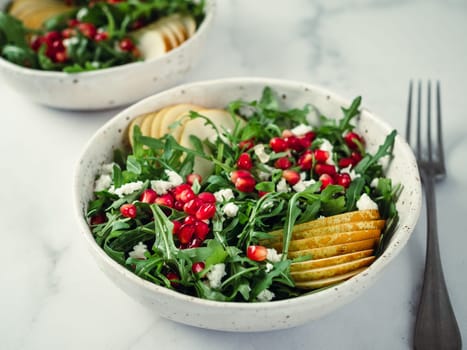 Fresh salad with arugula, pear, pomegranate and coconut crumble or cottage cheese. Two bowls with delicious summer fruit salad on marble table. Copy space for text