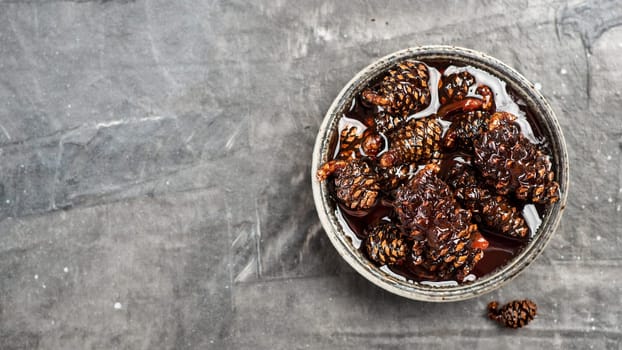 Delicious jam with baby pine cones in small bowl. Traditional Siberian dessert - young pine cones jam on gray textured background. Top view or flat lay. Copy space for text. Banner