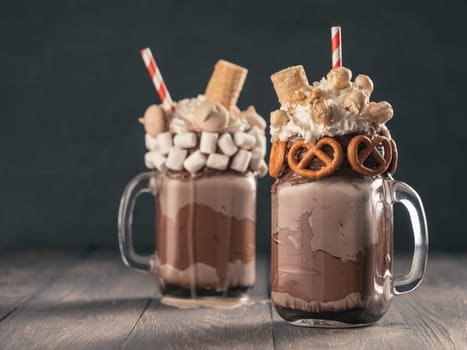 Close up view of two freakshake in mason jar on brown table. Freaked milkshake with chocolate, pretzel, marshmallow, popcorn and waffles. Trendy food concept - freak shake