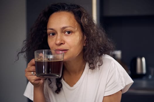 Beautiful woman holds a glass cup of freshly brewed americano, smiling looking at camera. Curly haired woman in white pajamas, enjoying her morning espresso coffee. Morning routine. People. Lifestyle