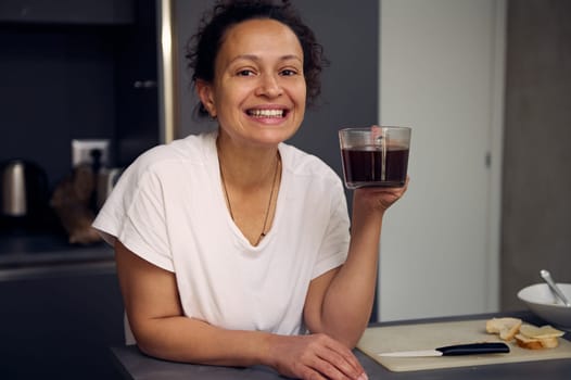 Happy smiling young woman in white pajama, standing at kitchen countertop and drinking coffee at home interior, smiling looking at the camera