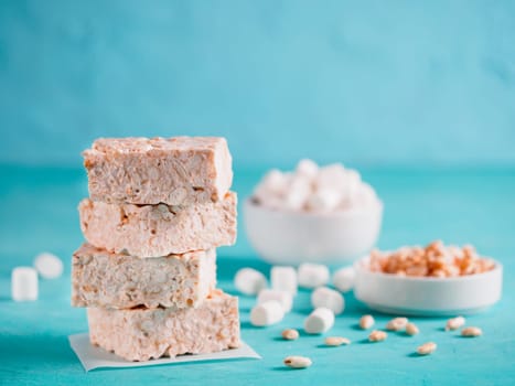 Homemade square bars of marshmallow and crispy rice with ingredients on azure blue background. Stack of American dessert with marshmallow and crispy rice. Copy space