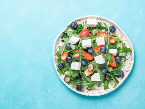 Salad with arugula, feta cheese and berries - strawberry, blueberry, in craft plate. Summer salad idea and recipe for healthy vegetarian lunch, dinner. Top view. Copy space. Colorful blue background.