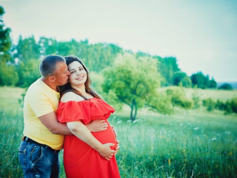 Pregnant beautiful woman in red dress and her husband with hands on belly outdoors. Man embraces from behind belly and kisses pregnant wife