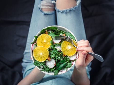 Woman in jeans at bed, holding vegan salad bowl with oranges, spinach,arugula,radish,nut.Top view.Vegan breakfast,vegetarian food,diet concept.Girl in jeans holding fork with knees and hands visible