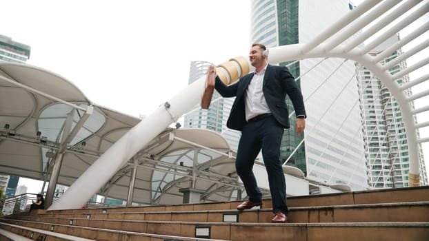 Happy businessman walking down stair and moving to music from headphone. Low angle camera view of smart manager listen song by using heaset and going back to home in urban city in lively mood. Urbane.