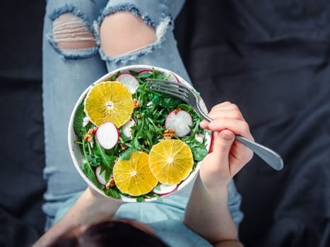 Woman in jeans at bed, holding vegan salad bowl with oranges, spinach,arugula,radish,nut.Top view.Vegan breakfast,vegetarian food,diet concept.Girl in jeans holding fork with knees and hands visible