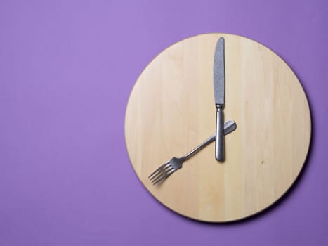 Intermittent fasting and skip breakfast concept - wooden round tray or trencher with cutlery as clock hands on lilac background. Eight hour feeding window concept or breakfast time concept. Copy space