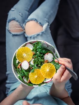 Woman in jeans at bed, holding vegan salad bowl with oranges, spinach,arugula,radish,nut.Top view.Vegan breakfast,vegetarian food,diet concept.Girl in jeans holding fork with knees and hands visible