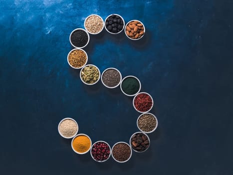 Above view of various superfoods in smal bowl in form S letter on dark blue background. Superfood as chia,spirulina,cocoa bean,goji, hemp, blueberry, quinoa, bee pollen,black sesame,turmeric. Top view