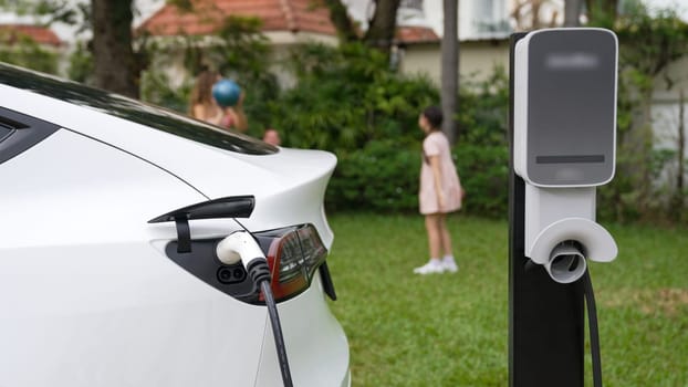 Electric vehicle recharge from home charging station on background of happy and playful family playing together. EV car using alternative and sustainable energy for better future Synchronos