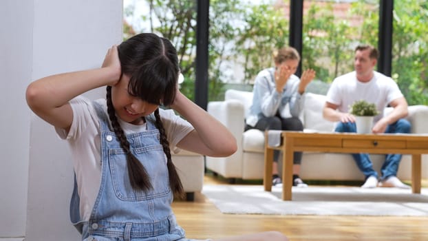 Stressed and unhappy young girl huddle in corner, cover her ears blocking sound of her parent arguing in background. Domestic violence at home and traumatic childhood develop to depression. Synchronos