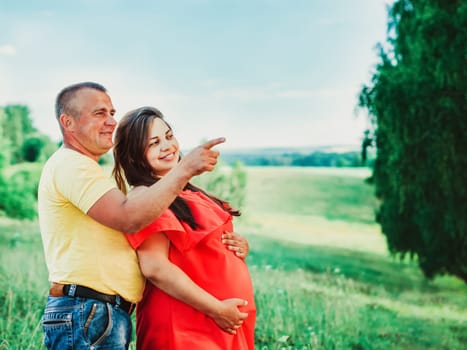 Pregnant beautiful woman in red dress and her husband with hands on belly outdoors. Man embraces from behind belly pregnant wife and shows his finger to the side