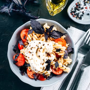 Tasty italian fusilli pasta with cherry, mozarella or buratta cheese and fresh basil. Dish with pasta on black concrete background. Top view. Copy space. Healthy food concept and recipe idea. Vertical