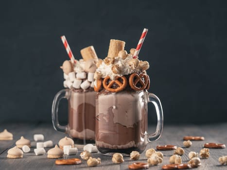 Close up view of two freakshake in mason jar on brown table. Freaked milkshake with chocolate, pretzel, marshmallow, popcorn and waffles. Trendy food concept - freak shake