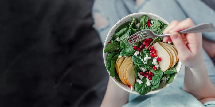 Woman in jeans holding vegan salad bowl with spinach, pear, pomegranate, cheese. Girl vegan in jeans holding fork with knees and hands visible. Banner. Copy space for text and design.
