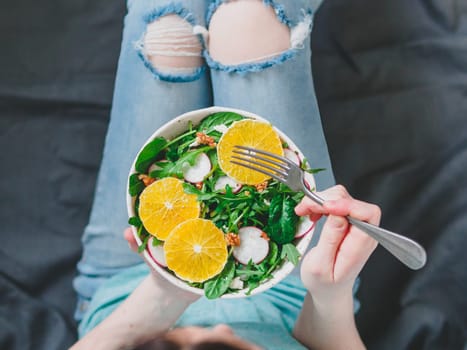 Woman in jeans at bed, holding vegan salad bowl with oranges, spinach,arugula,radish,nut.Top view.Vegan breakfast,vegetarian food,diet concept.Girl in jeans holding fork with knees and hands visible