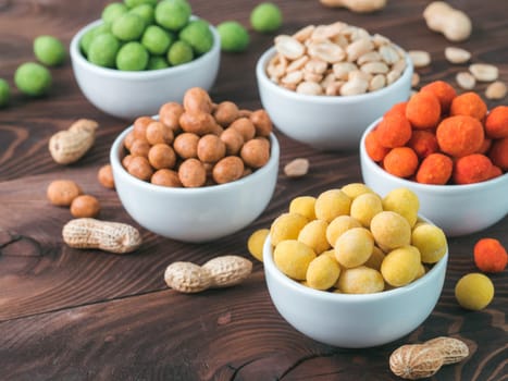 Assortment of different peanuts snack for beer, wine, party bar. Salted peanuts, peanuts in coconut glaze, green vasabi, red spicy chilli, yellow cheese glaze on brown wooden table. Copy space.