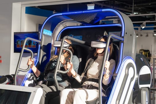 Moscow, Russia, March 14, 2024. Two woman is sitting in an electric blue virtual reality chair, wearing a headset. The machine combines engineering and fun in a display device