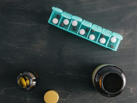 Top view of seven day pill box with pills. Green pill-box with pills visible. Open pill box and jars on dark wooden table. Copy space. Top view or flat lay.Healthy lifestyle and medical concept