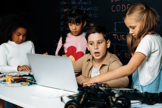 In classroom student in STEM class learning about coding robotics car. Brown shirt Schoolboy and white bib schoolgirl watching laptop and discussing. Other schoolgirls playing around funny. Erudition.