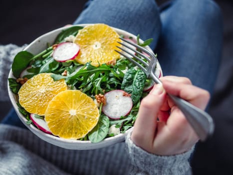 Woman in jeans at bed, holding vegan salad bowl with oranges, spinach,arugula,radish,nut.Top view.Vegan breakfast,vegetarian food,diet concept.Girl in jeans holding fork with knees and hands visible