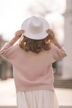 A woman wearing a white hat and a pink sweater stands in front of a building.