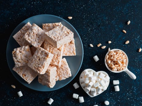 Homemade square bars of Marshmallow and crispy rice and ingredients on dark blue background. American dessert with marshmallow and crispy rice. Top view. Copy space