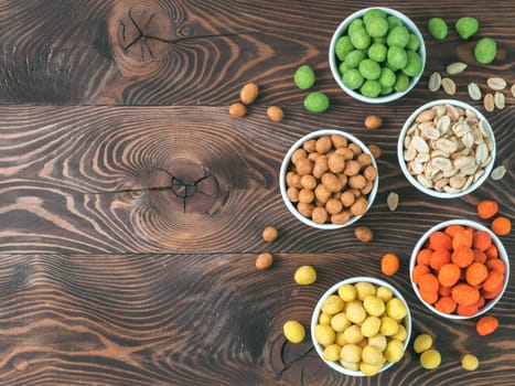 Assortment of different peanuts snack for beer, wine, party bar. Salted peanuts, peanuts in coconut glaze, green vasabi, red spicy chilli, yellow cheese glaze on brown wooden table. Copy space.