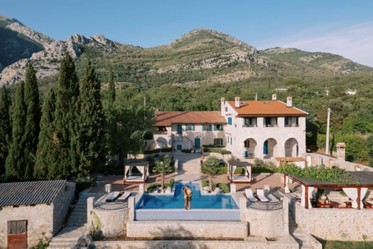 Man and woman stand hugging and kissing on the edge of a swimming pool near an old villa at the foot of the mountains. High quality photo