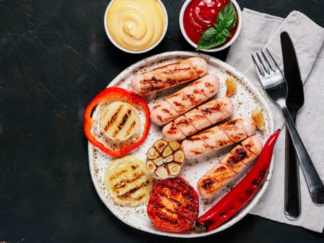 Top view of chicken homemade sausages with sauces ketchup and mustard on black background. Grilled sausages and grilled vegetables in craft trendy plate. Copy space.