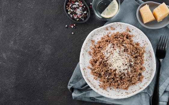 Buckwheat risotto with dried mushrooms served parmesan cheese in craft plate on black cement background. Gluten-free and vegetarian buckwheat recipe ideas. Copy space. Top view or flat-lay.