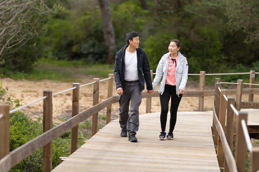 A man and woman are walking on a wooden bridge. The man is wearing a black jacket and the woman is wearing a pink jacket. They are holding hands and seem to be enjoying each other's company