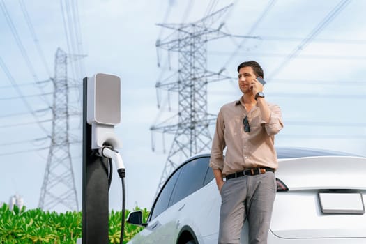 Man talking on the phone while recharge EV car battery at charging station connected to power grid tower electrical as electrical industry for eco friendly car utilization.Expedient