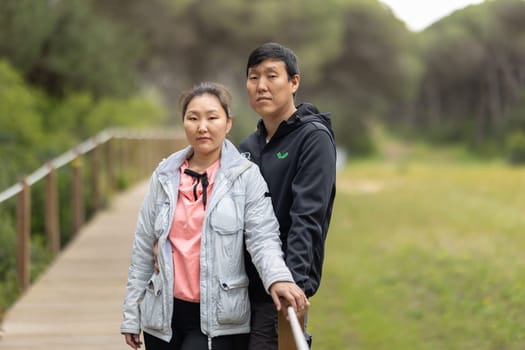 A man and woman are standing on a wooden bridge, with the man wearing a black jacket and the woman wearing a pink shirt