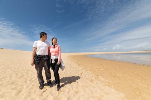 A couple is walking on a beach, enjoying the beautiful view of the ocean. The sky is clear and blue, and the sun is shining brightly. The couple is holding hands and smiling, creating a warm