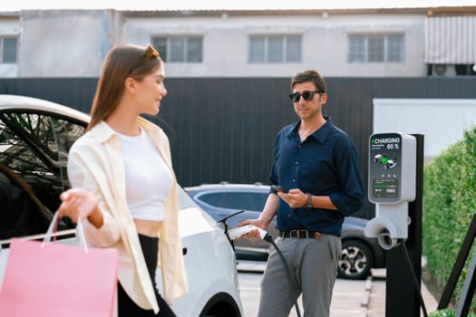 Young couple holding shopping bag and use smartphone to pay for electricity for recharging EV car battery from charging station at city mall parking lot. Happy couple go shopping by eco car. Expedient