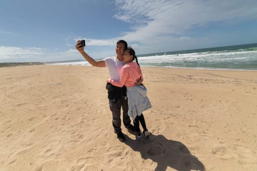 A man and woman are taking a selfie on a beach. The man is holding a cell phone and the woman is wearing a pink jacket. Scene is lighthearted and fun