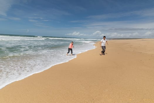 Two people are running on a beach with the ocean in the background. Scene is energetic and playful