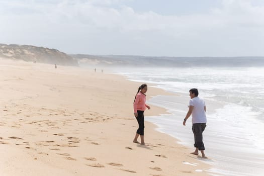 A man and a woman are walking on the beach. The woman is wearing a pink shirt. The beach is crowded with people